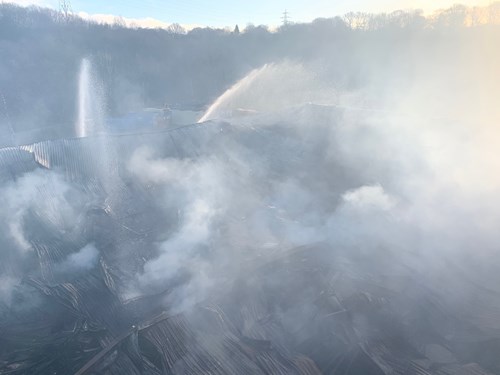 A smoke-filled scene showing water being sprayed over a large area, likely on smoldering materials, with thick gray smoke rising into the air, and trees and hills faintly visible in the background