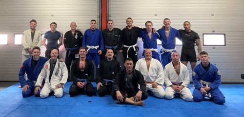 Group of men and women in Brazilian Jiu-Jitsu uniforms with various colored belts, sitting and standing on a blue mat in a training space