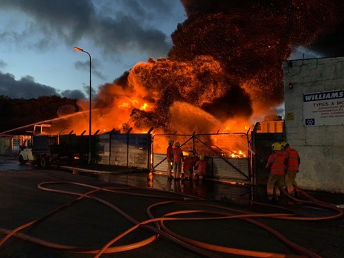 A large fire engulfs a building, producing thick black smoke and bright orange flames with several firefighters actively engaged in firefighting efforts