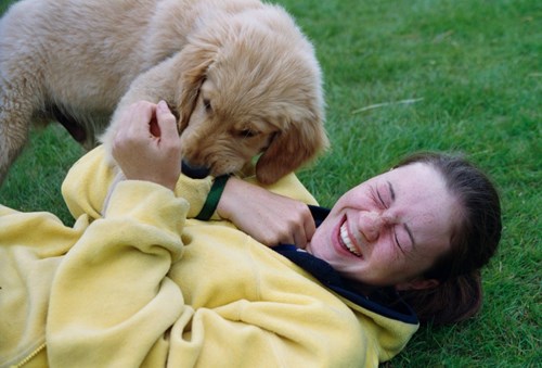 Vicky Peters lying on grass laughing in yellow fleece while a puppy is playing next to her