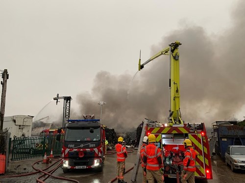 A picture of two fire engines with jets shooting water towards a large ploom of smoke and a number of firefighter around the vehicles