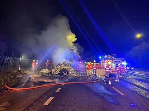 A fire scene on a road at night, with thick smoke rising from a burnt lorry and multiple firefighters in high-visibility jackets working to extinguish the flames