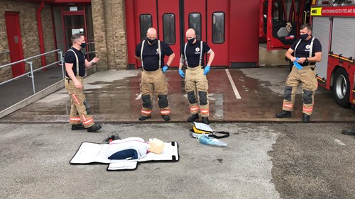 Five firefighters in training uniforms perform CPR on a training dummy outside a fire station