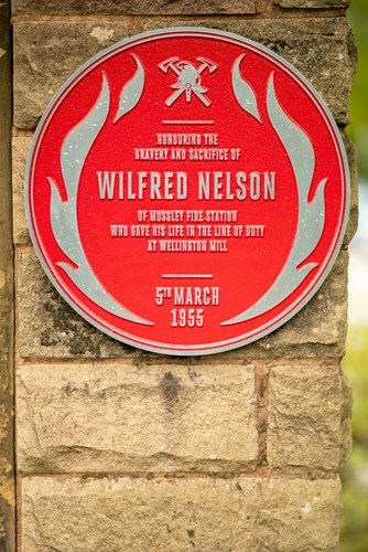 A circular red plaque mounted on a stone wall, honoring Wilfred Nelson, a firefighter from Mossley Fire Station. It reads: 'Honouring the bravery and sacrifice of Wilfred Nelson who gave his life in the line of duty at Wellington Mill, 5th March 1955.