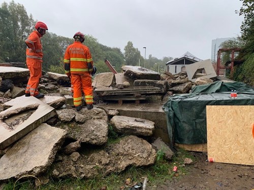 Two workers in bright orange safety gear survey a scene of debris consisting of broken concrete slabs and rubble, with a partially visible building in the background