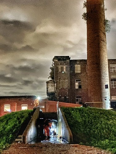 A search and rescue exercise taking place in a dimly lit industrial area, featuring a dog and rescue personnel