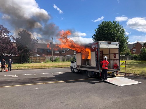 Image shows flames licking out of kitchen safety unit demonstrating how a cooking fire can break out. Firefighter stood to right of image in orange uniform and white helmet.