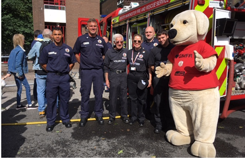 Six firefighters and a dog mascot pose with a fire engine at an Oldham community event