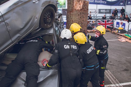 GMFRS RTC team competing in a 20-minute challenge to extricate a casualty out of a damaged vehicle, partially crushed under another car and against a tree, in a simulated accident. the team are wearing black uniform with yellow helmets.