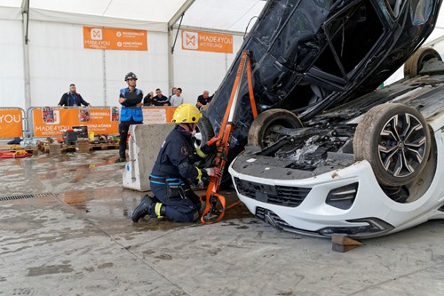 A rescue worker in a yellow helmet kneels beside an overturned car, using equipment to stabilize or lift the vehicle during a complex scenario in a 30-minute challenge