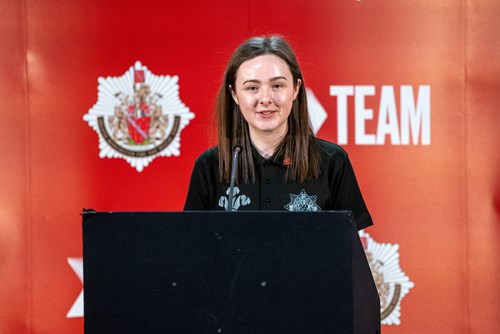An image of a young woman standing at a lectern in front of a red GMFRS banner