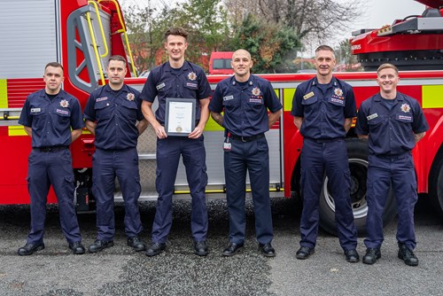 Six firefighters stand in a row in front of a fire engine. The firefighter in the centre holds a certificate.