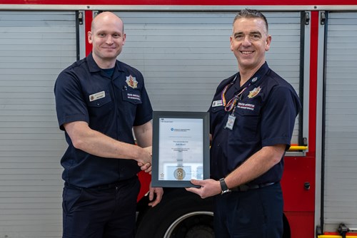 Two smiling firefighters stand in front of a fire engine inside a fire station.