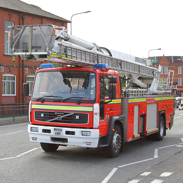 A red and white fire truck with a raised ladder driving on a street near buildings, displaying fire service markings