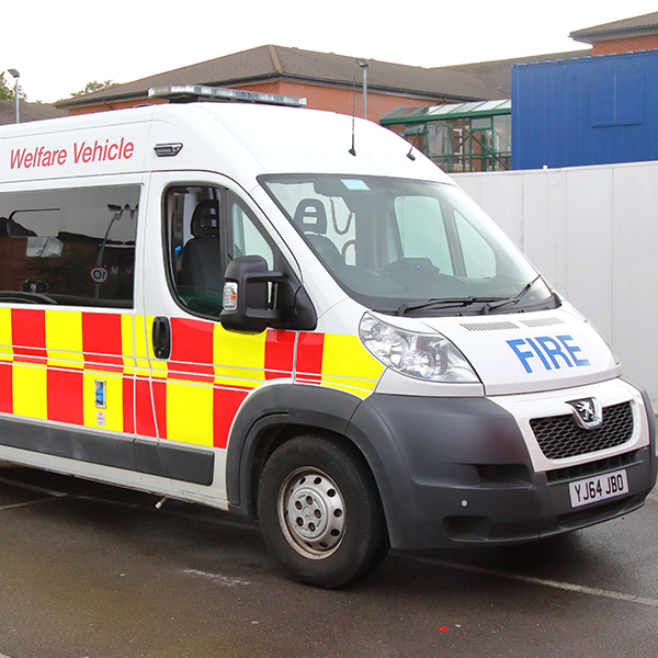 A welfare vehicle used by fire services, parked on a road, with a building and a blue structure in the background