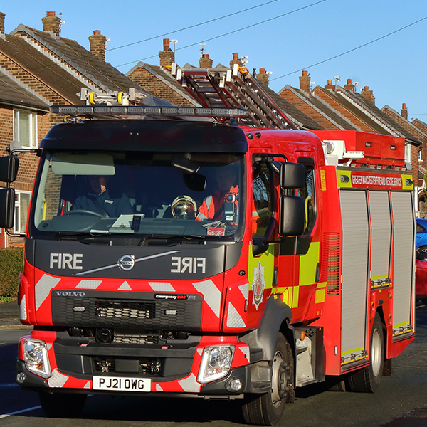 A red fire truck with "FIRE" displayed on the front is parked on a street lined with residential houses. The truck has a ladder on top and various emergency lights visible. The vehicle is actively engaged, with a fire service personnel visible in the driver's seat, wearing a helmet