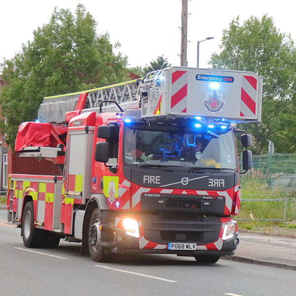 A red and white fire truck from Stretford Community Fire Station with a ladder on top, equipped with flashing blue lights, driving down a street with trees and residential buildings in the background