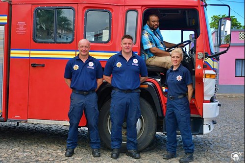 Three firefighters stand in front of a fire engine, and another man sits in the driver's seat of the engine