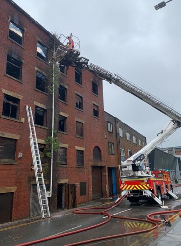 A firefighter in an orange suit uses a ladder to access a burned brick building with broken windows, while a fire truck and hoses are nearby