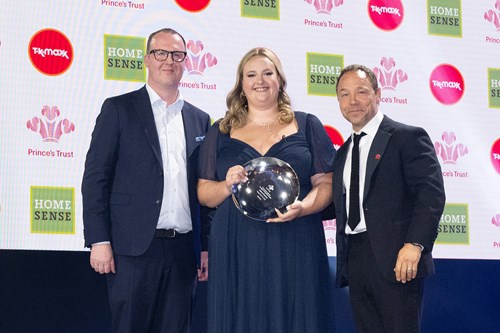 Joanna, flanked by two men in suits, holds her award in front of a screen displaying the Prince's Trust logo