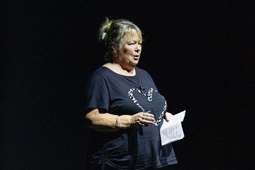 A woman with shoulder-length blonde hair speaks to an audience on stage, wearing a black shirt. She gestures with one hand while holding a piece of paper in the other.