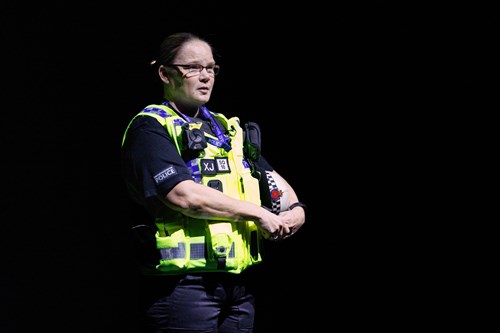 GMP police constable Nicola Heap wearing a high-visibility vest stands in front of a dark background, addressing an audience
