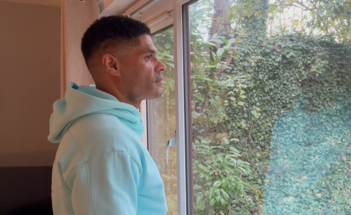 A man in a light blue hoodie stands next to a window, looking out at a greenery-filled outdoor scene. The man has a thoughtful expression.