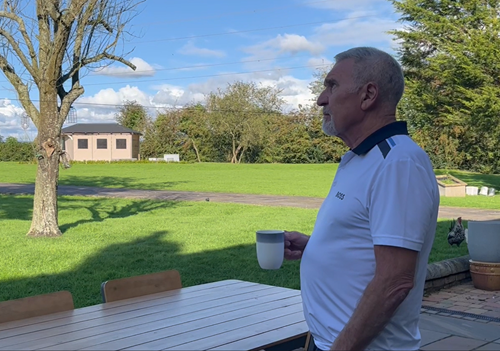 A man stands outside in a casual white shirt, holding a cup, while looking thoughtfully at a green landscape with trees and a clear blue sky
