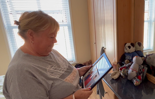 A woman smiling and looking at a photo of late son Colin Hornsby who passed away in a crash. She stands in a bright room with large windows, surrounded by stuffed animals on a desk.