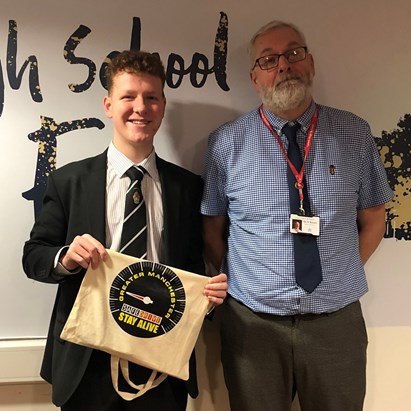 David Baker a young male student in a school uniform, smiling and holding a Drive Safe tote bag, stands next to a male teacher in a blue shirt and tie in a school environment