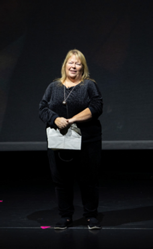 Anne Marie Hornsby stood on stage against black background looking out at audience and holding piece of paper