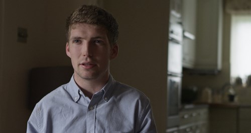 Joe Robinson a young man with light brown hair seated in a soft-focus kitchen area, wearing a light blue collared shirt and looking slightly off-camera