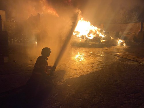 A firefighter kneels on the ground, aiming a hose at a large fire engulfing a pile of scrap materials in a dark, smoky environment