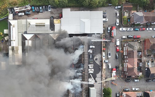 Aerial view of a large fire at a commercial unit in Levenshulme, with thick smoke rising. Firefighters and equipment are present, along with emergency vehicles on nearby streets