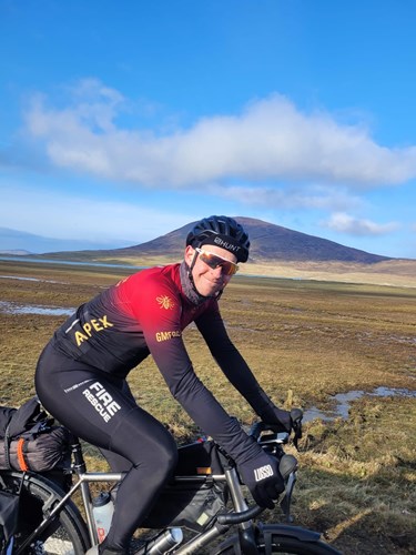 A GMFRS cyclist wearing a black and red cycling jersey and a helmet is smiling while sitting on their bicycle. The background features a scenic landscape with a mountain, open fields, and clear blue skies
