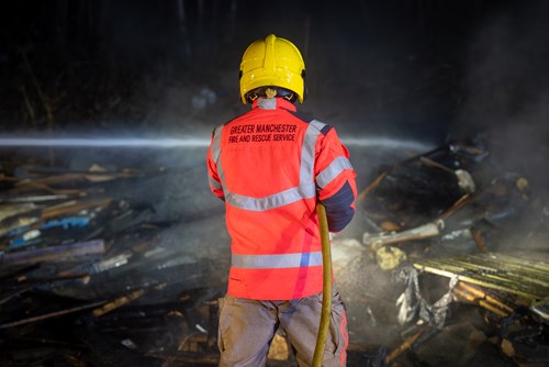 Firefighter in bright orange and grey uniform directs water onto debris during a controlled fire