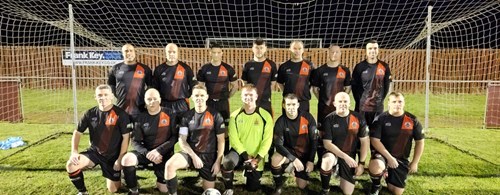 A veteran football team in black uniforms with red accents pose in front of a goal post on a grassy field