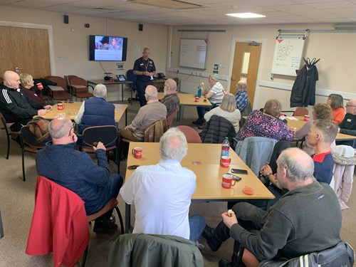 Living well group members sat around tables with refreshments listening to a speaker from the fire service stood by a screen