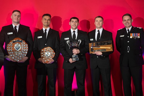 Five individuals in formal black uniforms stand side by side against a red backdrop. They are holding various awards and trophies, including a large shield, a cup, and a box trophy. The expressions on their faces are proud. One person, wearing a uniform with medals, is positioned to the far right, slightly apart from the others