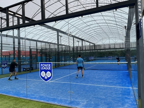 Padel court at The Power League Manchester. The court surface is blue, and a net runs across the centre line. There are four players using the court.